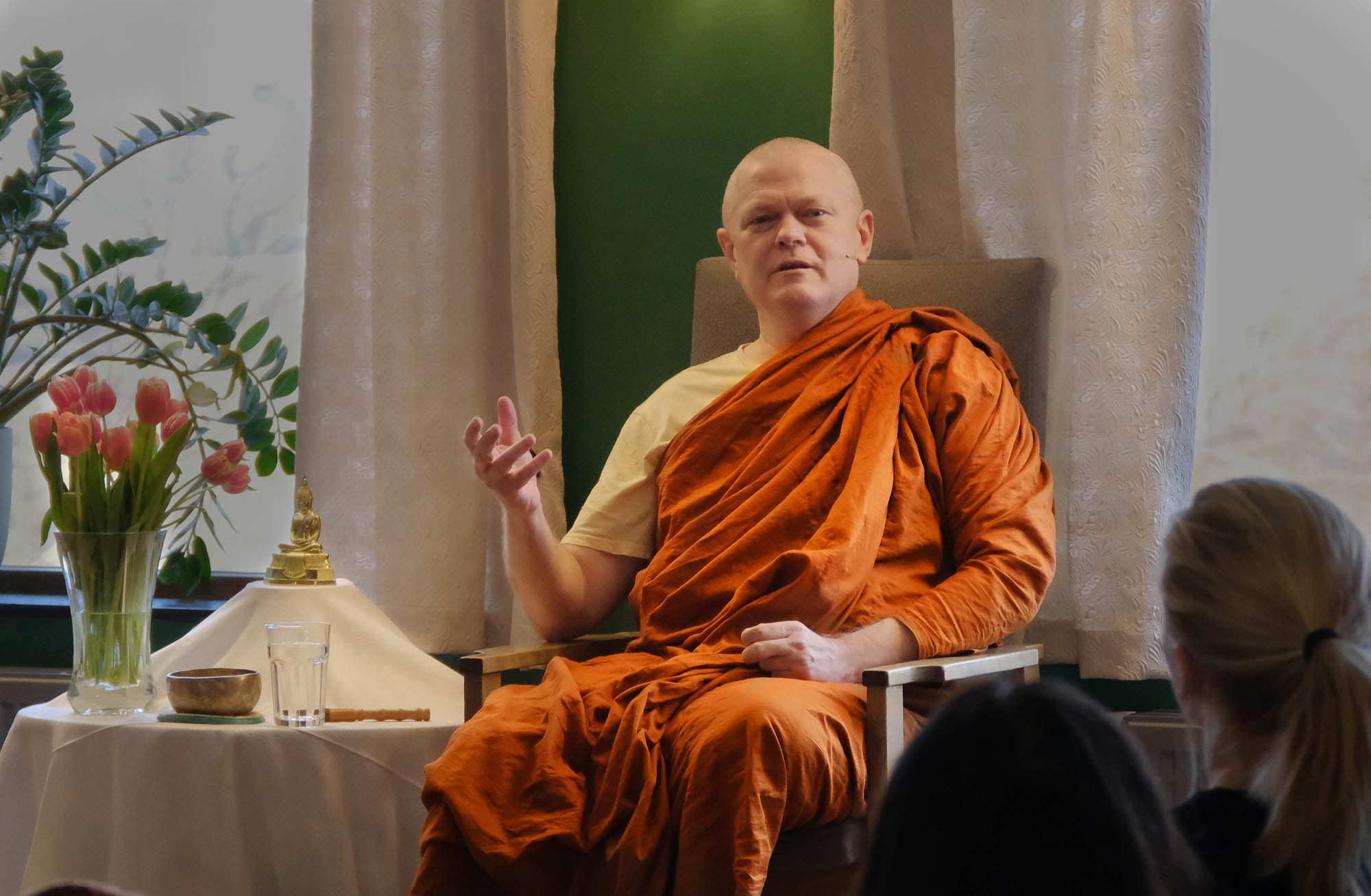 Bhante Sujato in monks robes, seated in a chair, next to a small shrine, teaching meditation to a group of people